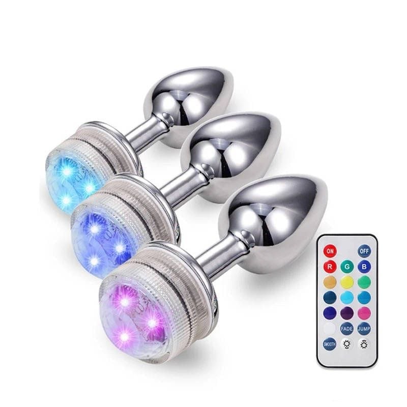 Three polished stainless steel butt plugs with illuminated LED gems in blue, purple, and pink, displayed with a wireless remote control