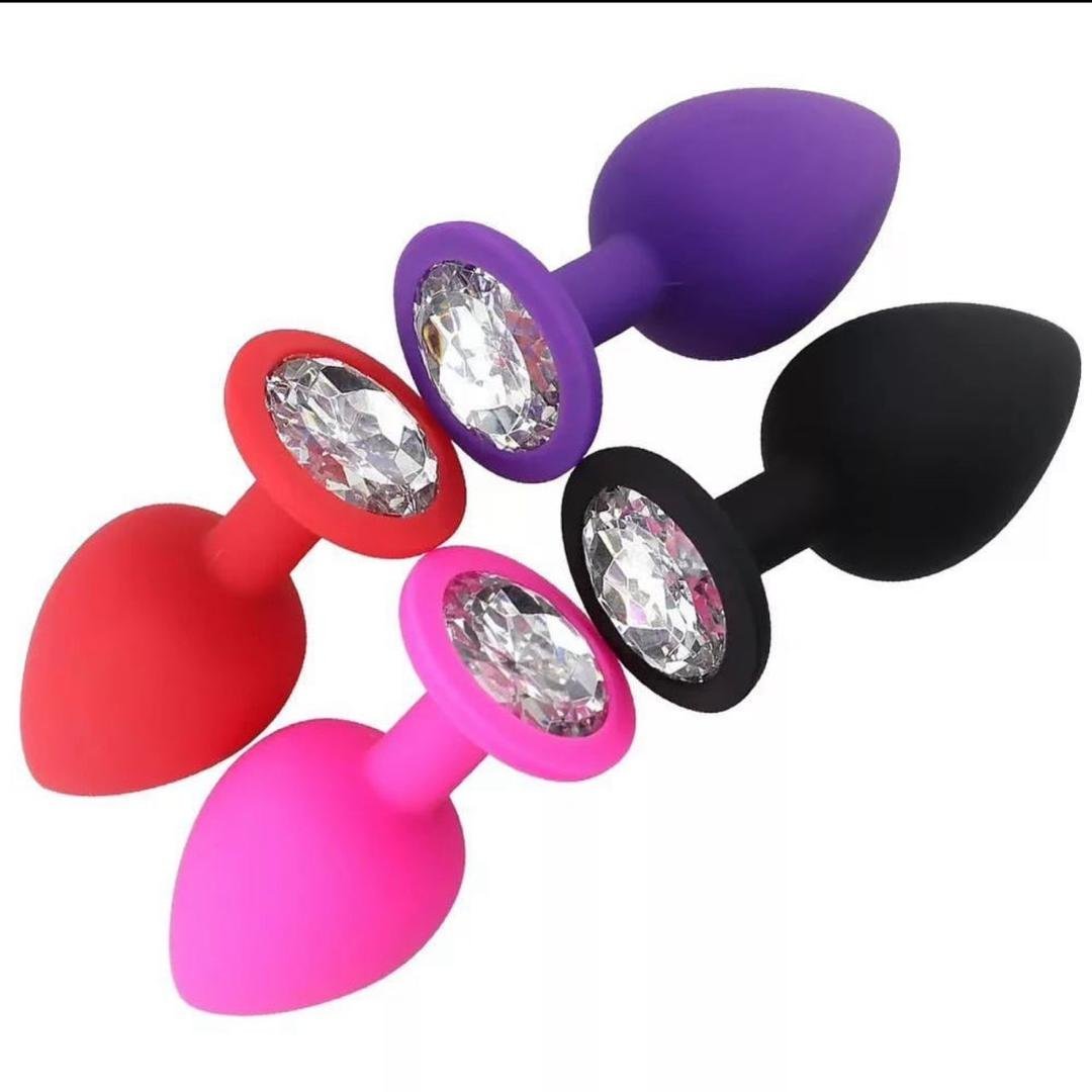 Four colorful silicone butt plugs with jeweled bases in red, pink, purple, and black arranged in a diagonal line